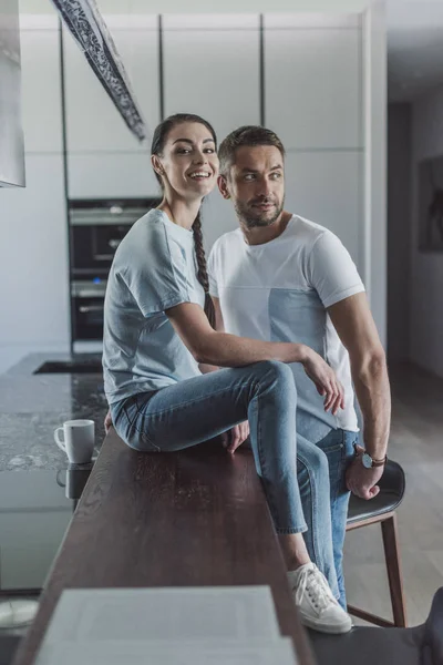 Feliz joven sentada en el mostrador de la cocina mientras su novio de pie cerca de casa — Stock Photo