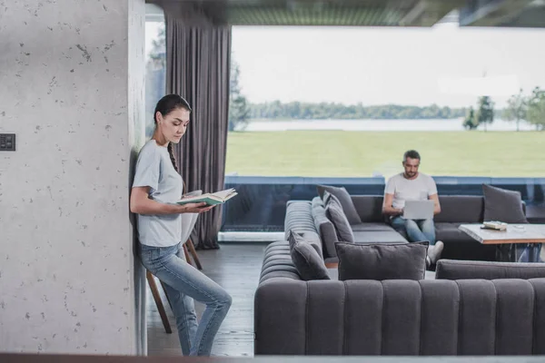 Side view of woman reading book while her boyfriend using laptop behind at home — Stock Photo