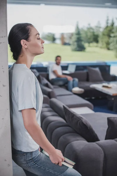 Вид збоку жінки з закритими очима тримає книгу, а її хлопець використовує ноутбук позаду вдома — стокове фото