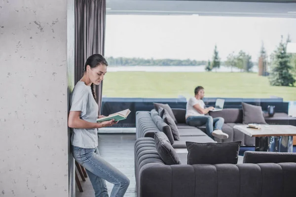 Vista lateral de la mujer leyendo libro mientras que su novio usando el ordenador portátil detrás en casa - foto de stock