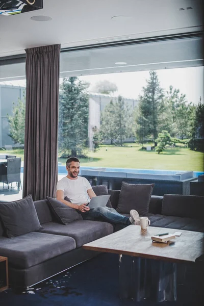 Souriant adulte mâle indépendant travaillant avec ordinateur portable sur canapé à la maison — Photo de stock