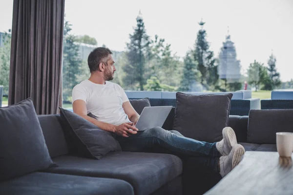 Adult male freelancer working with laptop and looking away on sofa at home — Stock Photo