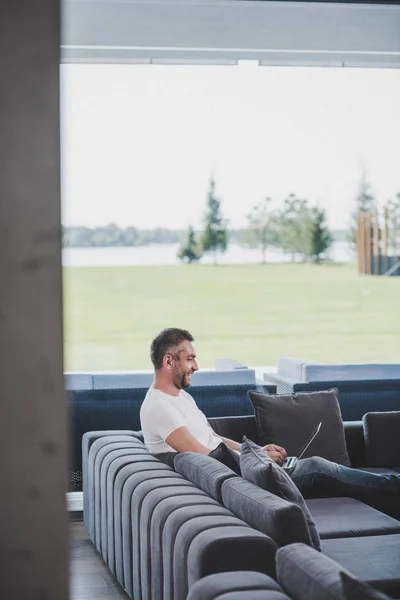 Side view of smiling adult man using laptop on couch at home — Stock Photo