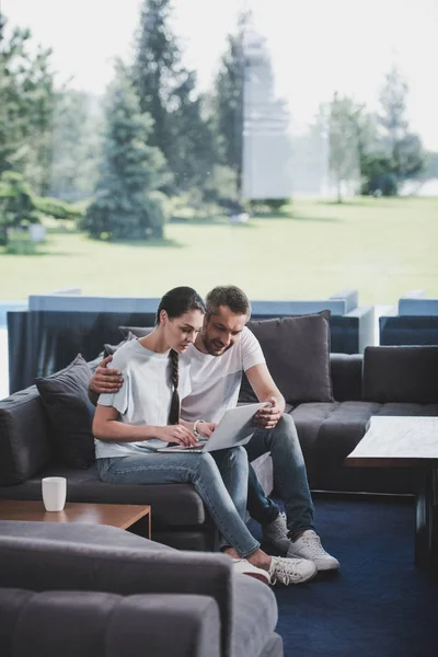 Adult man embracing girlfriend and teaching her using laptop on couch at home — Stock Photo