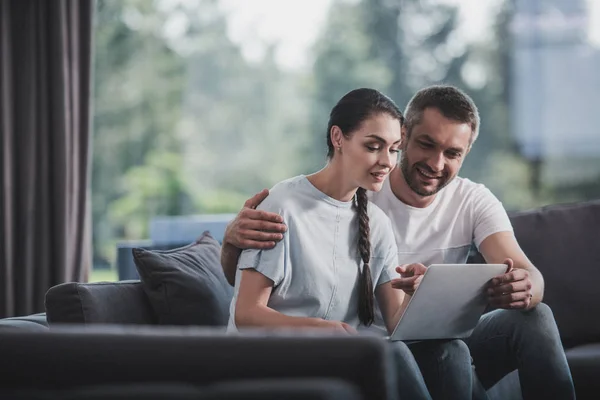 Feliz hombre abrazando novia y enseñándole a usar el ordenador portátil en el sofá en casa - foto de stock