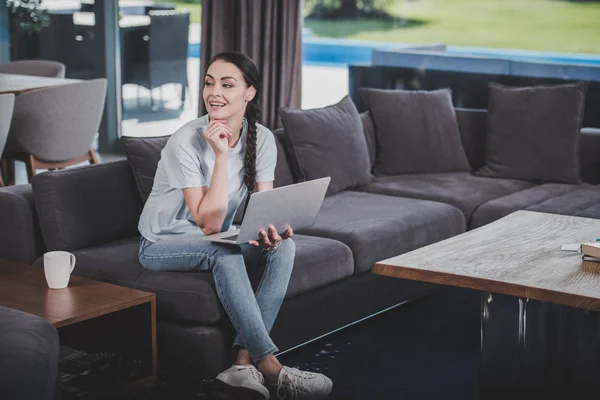 Sonriente freelancer femenina trabajando con laptop en sofá en casa - foto de stock