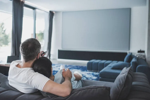 Vista posteriore dell'uomo adulto con telecomando che abbraccia la fidanzata e guarda la tv sul divano a casa — Foto stock