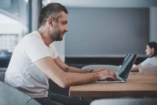 Vista lateral del freelancer masculino sonriente usando el ordenador portátil con la pantalla en blanco en casa - foto de stock