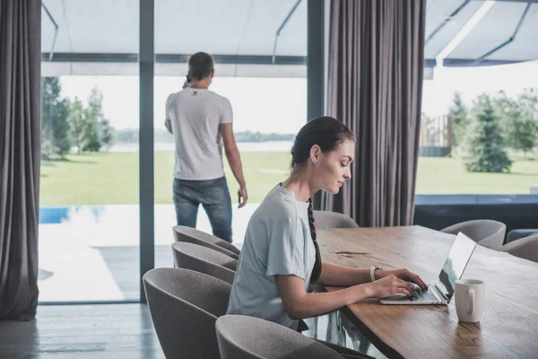 Vista lateral da jovem mulher usando laptop na mesa, enquanto seu namorado falando no smartphone em casa — Fotografia de Stock