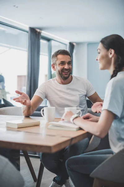 Homem adulto feliz gesticulando e conversando com a namorada à mesa com xícaras de café e livros em casa — Fotografia de Stock