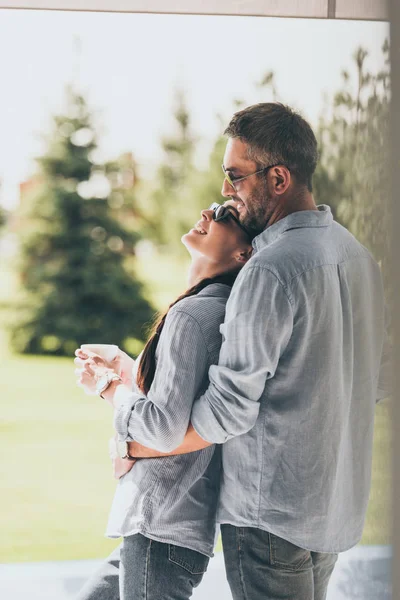 Enfoque selectivo del hombre en gafas de sol abrazando novia con taza de café al aire libre - foto de stock