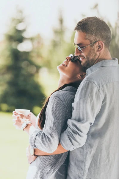 Vista lateral de hombre feliz en gafas de sol abrazando novia con taza de café al aire libre - foto de stock