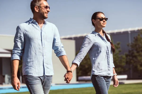 Elegante pareja en gafas de sol cogidas de la mano y caminando sobre césped verde en casa de campo - foto de stock