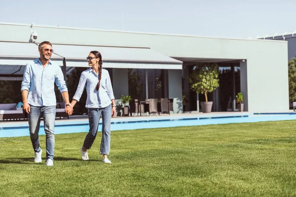 Sonriente pareja cogida de la mano y caminando sobre césped verde cerca de la piscina en la casa de campo - foto de stock