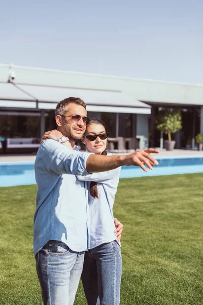 Adult man in sunglasses pointing by finger to girlfriend standing near on green lawn at country house — Stock Photo