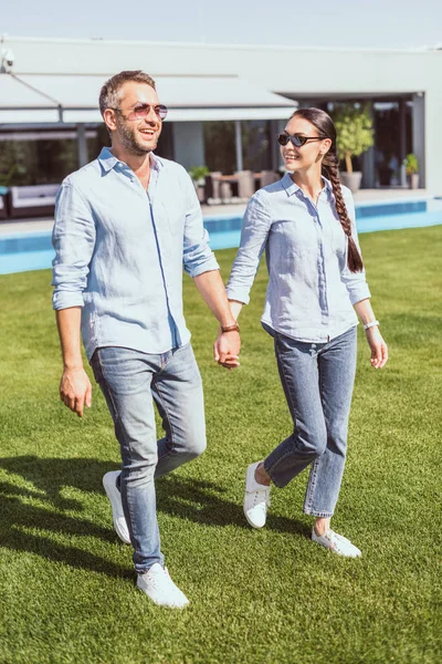 Couple heureux dans des lunettes de soleil tenant la main et marchant sur la pelouse verte à la maison de campagne — Photo de stock