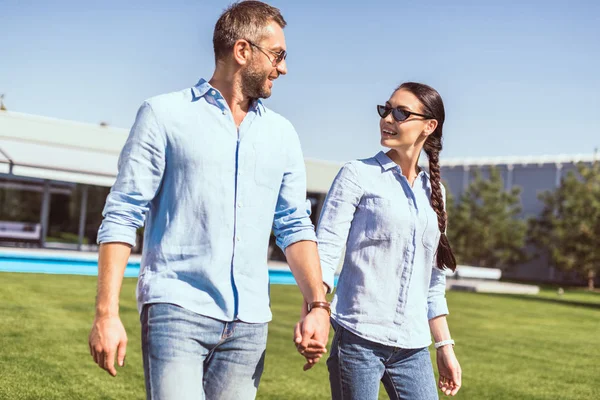 Couple dans des lunettes de soleil tenant la main et se regardant sur la pelouse verte à la maison de campagne — Photo de stock