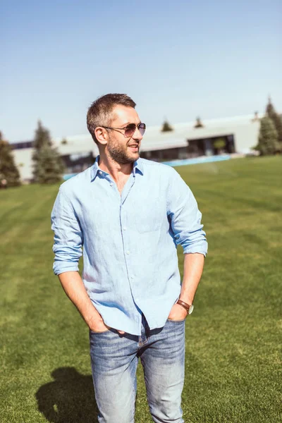 Hombre guapo feliz en gafas de sol caminando con las manos en los bolsillos en la hierba cerca de casa de campo - foto de stock