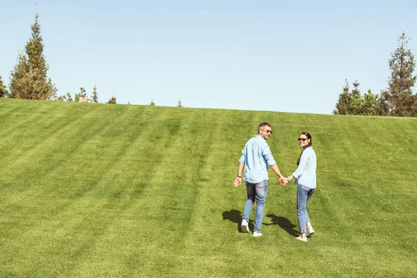 Casal elegante em óculos de sol de mãos dadas e olhando para a câmera na colina gramada ao ar livre — Fotografia de Stock