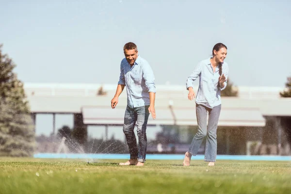 Casal feliz se divertindo com rega automática no gramado verde perto da casa de campo — Fotografia de Stock