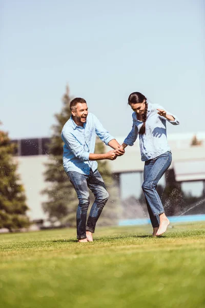 Adulto ridendo uomo tenendo la mano della fidanzata e divertirsi con irrigazione automatica sul prato verde vicino casa di campagna — Foto stock