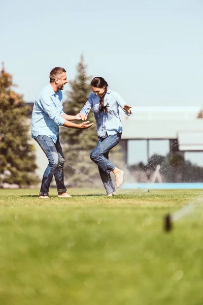 Ridere coppia divertirsi con irrigazione automatica sul prato verde vicino casa di campagna — Foto stock