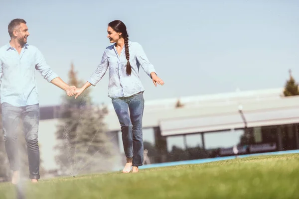 Happy couple holding hands and having fun with automatic watering on grass near country house — Stock Photo