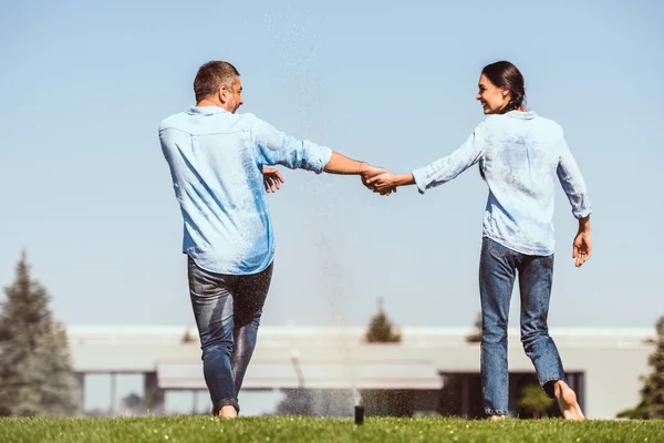 Вид сзади пары, держащейся за руки и танцующей под автоматическим поливом на зеленой лужайке возле загородного дома — стоковое фото