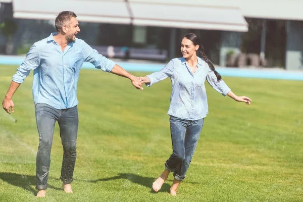 Sonriente pareja cogida de la mano y divertirse con riego automático en el césped verde cerca de la casa de campo - foto de stock