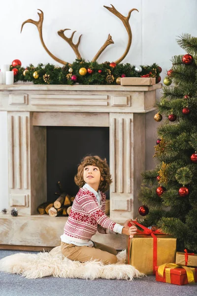 Adorable niño feliz sentado en el suelo con cajas de regalo de Navidad y mirando hacia arriba - foto de stock