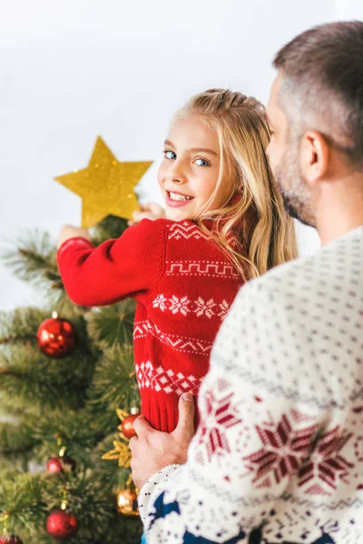 Bonito jovem pai carregando sua filha enquanto ela decorando árvore de natal — Fotografia de Stock