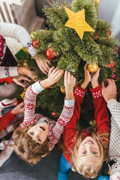 Vista de ángulo alto de los niños decorando el árbol de Navidad con la familia y mirando a la cámara - foto de stock