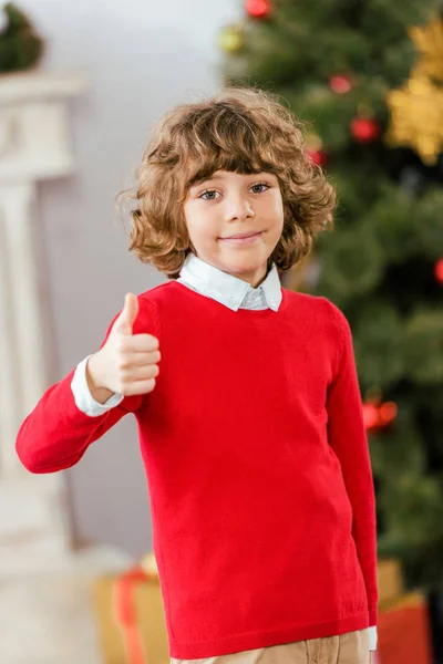 Adorável criança feliz mostrando polegar para cima com árvore de natal borrada no fundo — Fotografia de Stock
