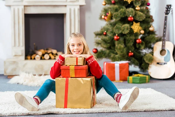 Adorable happy child leaning on stack of christmas gift boxes while sitting on floor — Stock Photo