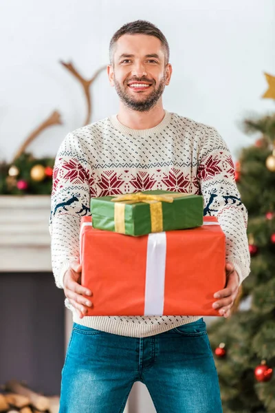 Щасливий бородатий чоловік у різдвяному светрі з подарунковими коробками дивиться на камеру — стокове фото