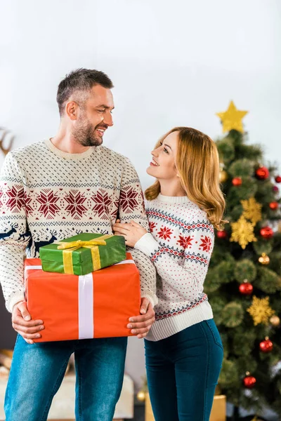 Glückliches Paar im Weihnachtspullover mit Geschenkschachteln, die sich anschauen — Stockfoto