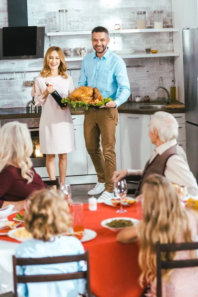 Молодой человек и женщина несут вино и индейку на ужин в честь Дня благодарения с семьей дома — стоковое фото