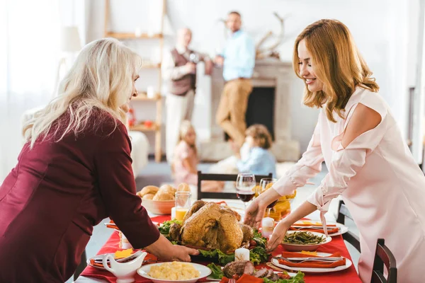 Sorridenti donne giovani e anziane che servono la tavola del giorno del ringraziamento mentre uomini e bambini in piedi offuscati sullo sfondo — Foto stock
