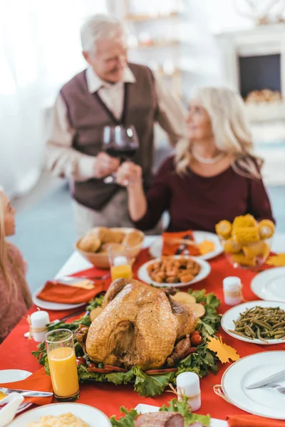 Vários deliciosos alimentos na mesa em primeiro plano com copos de vinho de casal sênior durante a celeração de ação de graças com neta — Fotografia de Stock