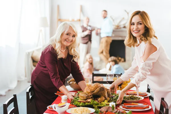 Happy young and senior women serving thanksgiving day table while men and kids standing blurred on background — Stock Photo