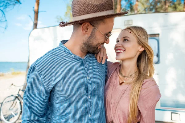 Feliz joven pareja abrazando cerca de caravana en el bosque - foto de stock