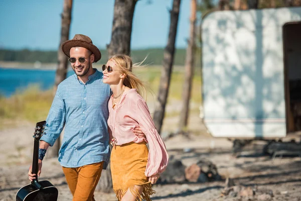 Bella coppia hippie in occhiali da sole abbracciare e camminare con la chitarra vicino camper — Foto stock