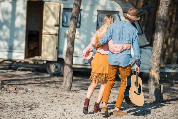 Rückansicht von Hippie-Paar, das sich umarmt und mit Gitarre in der Nähe von Wohnwagen spaziert — Stockfoto