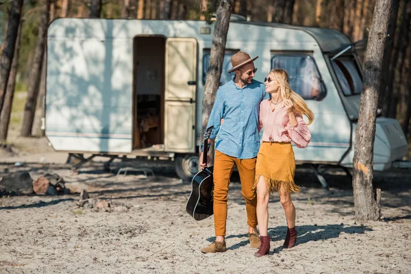 Beautiful couple hugging and walking with acoustic guitar near campervan in forest — Stock Photo