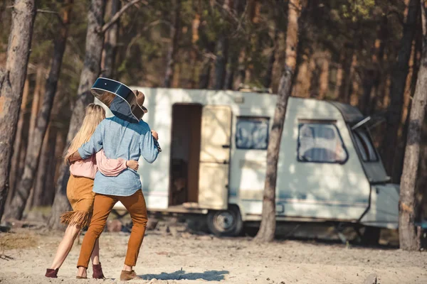 Vista trasera de pareja abrazando y caminando con guitarra acústica a autocaravana en bosque - foto de stock