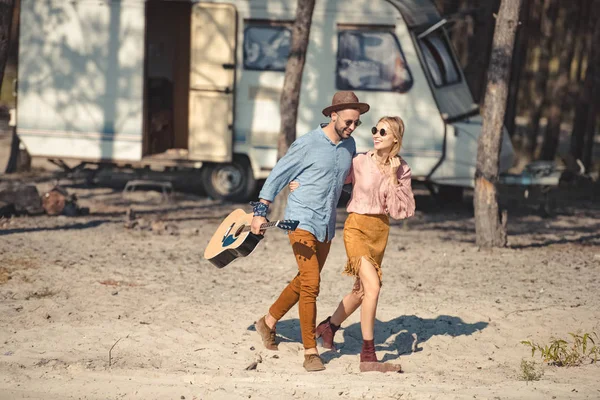Schönes Paar mit Sonnenbrille umarmt und spaziert mit Akustikgitarre in der Nähe von Wohnwagen im Wald — Stockfoto