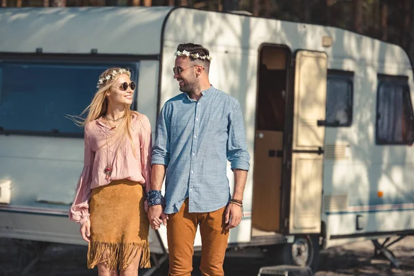 Pareja hippie mirándose mientras se toman de la mano y posan cerca de campervan - foto de stock