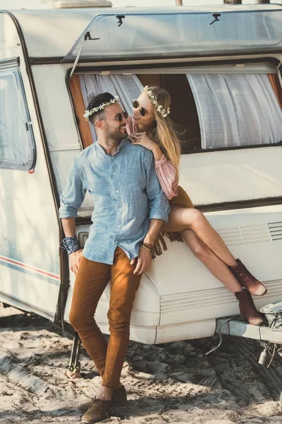 Hippie couple in wreaths and sunglasses looking at each other and sitting on trailer — Stock Photo