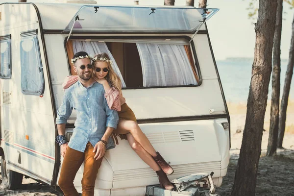 Hippie couple in wreaths and sunglasses hugging and sitting on trailer near sea — Stock Photo