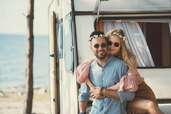 Smiling hippie couple in wreaths and sunglasses embracing and sitting on campervan — Stock Photo